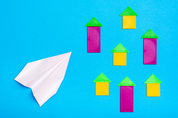 White paper plane flies over colored figures of houses on a blue cardboard background. Top view. Plane crash danger concept