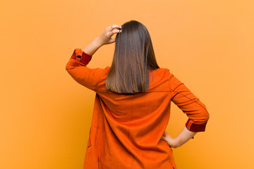 young pretty woman feeling clueless and confused, thinking a solution, with hand on hip and other on head, rear view isolated against orange wall