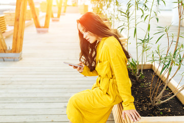 Happy girl using a smart phone in a city park sitting in yellow dress with long hair