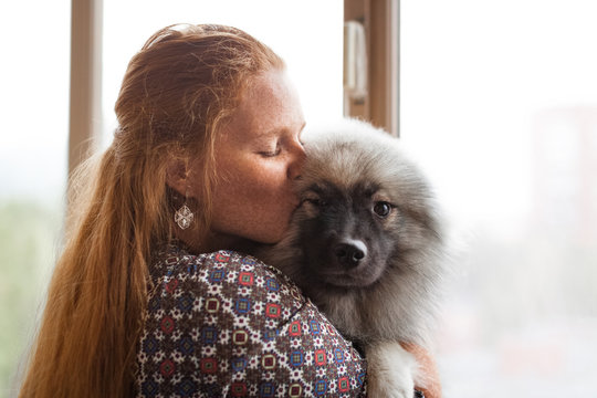 Young red-haired woman stands by the window with a Keeskhond puppy in her arms and kisses it