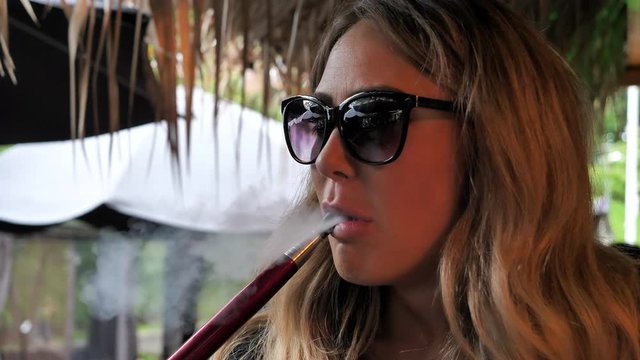 Blonde girl with attitude wearing sunglasses blowing out smoke from water pipe. Close up.