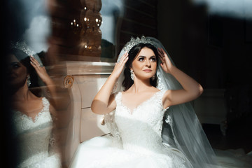 Naklejka premium Morning of the bride. Wedding photography. Beautiful bride portrait wedding makeup hairstyle, gorgeous young woman in white dress at home. The bride touches the crown.