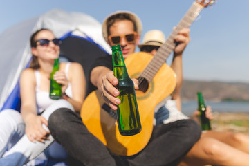 Guitar player showing bottle of beer while having a great time with his friends during camping at the beach.