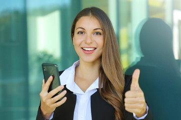 Business woman using mobile phone shows thumb up outdoors.