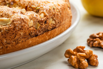 A large pie with apples, chopped walnuts and cinnamon lies on a white plate on a light background next to walnuts and a yellow apple
