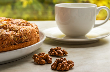 Pie with apples, chopped walnuts and cinnamon lies on a white plate on a light background next to walnuts and a white cup on a saucer