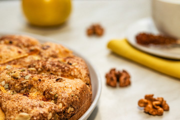 Pie with apples, chopped walnuts and cinnamon lies on a white plate on a light background next to walnuts, a yellow apple and a white cup and saucer on a yellow napkin