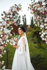 Beautiful fiancee in white wedding dress with bouquet stand near the wedding arch of flowers. Portrait of a cute bride with a bouquet in the scenery. Wedding day. Fashion bride.