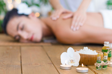 Obraz na płótnie Canvas Closeup burning candles, bottles with aroma oil, bowl with salt. Young pleased woman is getting thai massage, therapy. Brunette girl is lying on couch in spa ayurveda salon. Relax and health care.