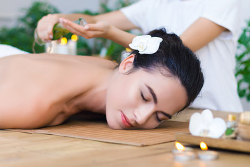 Obraz na płótnie Canvas Young pleased woman is getting thai massage, therapy. Female master is pouring massaging oil in palms. Brunette girl is lying on couch in light spa ayurveda salon. Relax and health care concept.