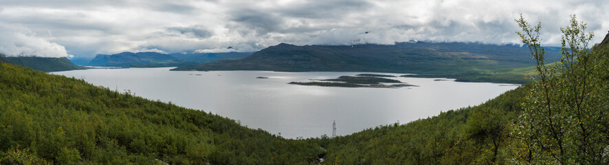 Panoramic landscape of Akkajaure lake at Kungsleden hiking trail, green mountains and birch tree forest in mist and clouds