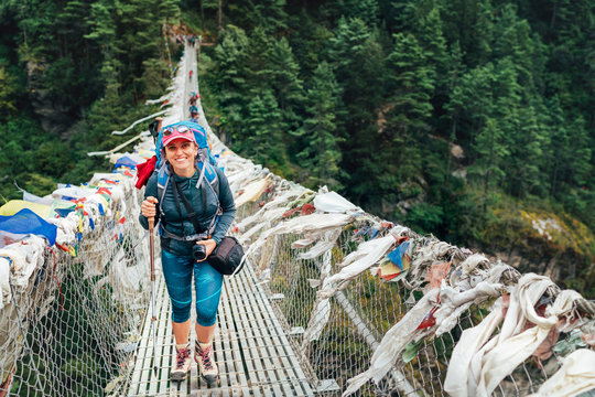 Young smiling female photographer crossing canyon over Suspension Bridge decorated with multicolored Tibetan Prayer flags hinged over gorge.  Everest Base Camp route, Sagarmatha National Park, Nepal.