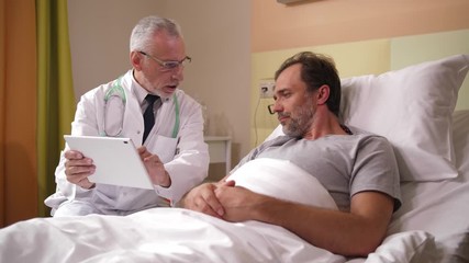 Closeup of aging physician in white robe sitting next to sick male lying in hospital ward and showing positive tomography results. Elderly practitioner and adult patient watching brain scans on tablet