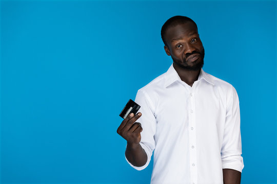 playful looking forward afroamerican man in white shirt is holding credit card in the right hand on the blue background
