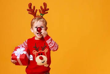 happy funny child boy in red Christmas reindeer costume with gift on yellow   background