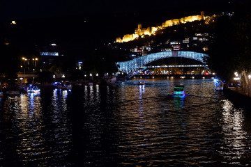 Fototapeta premium The Bridge of Peace is a bow-shaped pedestrian bridge, a steal and glass construction illuminated with numerous LEDs, over the Kura River in downtown Tbilisi, capital of Georgia. Night photo.