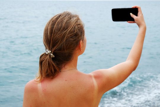 Young girl in a swimsuit makes a selfie with a smartphone on a beach. View to the azure sea water and sand, romance and vacation concept. Place for an inscription.