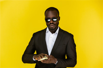 bearded serious young afroamerican guy is holding dollars in both hands, wearing sunglasses and...