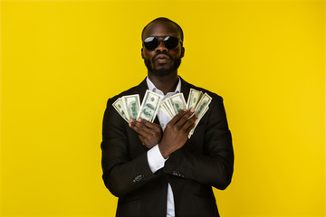 bearded luxury young afroamerican guy is holding lots of money in both hands in sunglasses and black suit on the yellow background