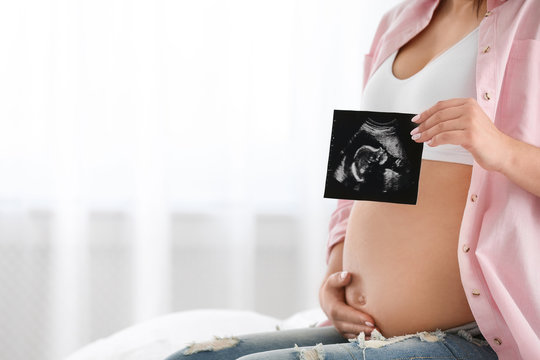 Pregnant woman with ultrasound picture indoors, closeup. Space for text