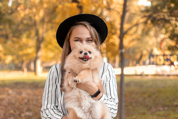 A girl of European appearance in a black hat with blond hair is holding a dog of the breed Pomeranian Spitz of red color on the background of autumn leaves. Emotion of joy.
