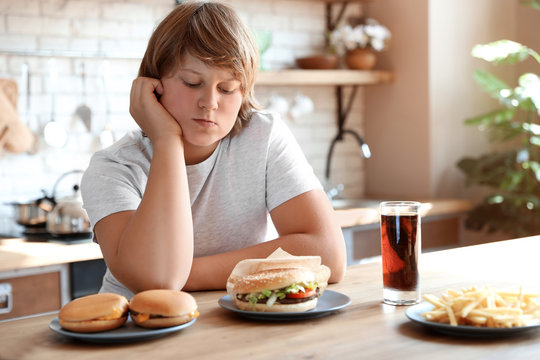 Overweight boy at table with fast food in kitchen
