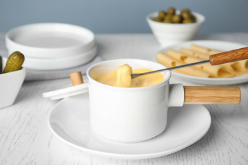 Pot of tasty cheese fondue and fork with bread on white wooden table