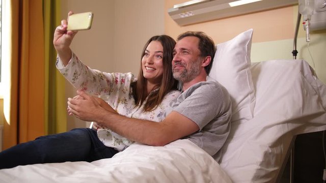 Close-up of happy married couple posing for selfie photo on cellphone in hospital ward. Pretty woman encouraging recovering husband during visit in clinic. Joy, love, togetherness, support, care