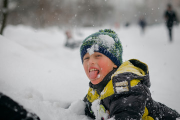 Fototapeta na wymiar boy sits in snowdrift and shows tongue. child was thrown with snow and thrown into snowdrift. boy walks on street after snowfall. Active healthy lifestyle