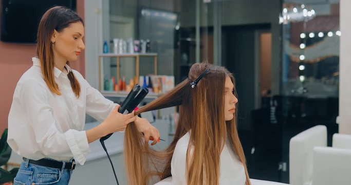 Professional hair stylist separates a strand of hair and straightens it. Beautiful female model on procedure in modern beauty salon.