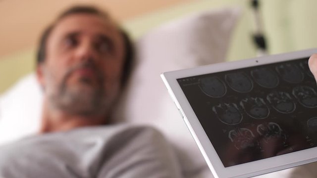 Close-up of male physician hands holding tablet with results of MRI with patient lying in bed blurred on background. Practitioner telling ill man about tomography examination