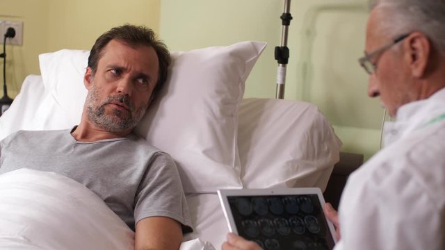 Close-up of concerned patient listening to elderly doctor telling disappointing diagnosis based on results of MRI. Aging practitioner talking to unhappy male while watching brain scans on tablet pc