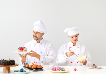 Young confectioners cooking tasty desserts against light background