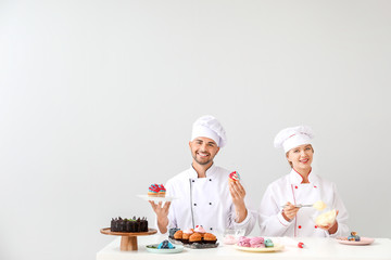 Young confectioners cooking tasty desserts against light background