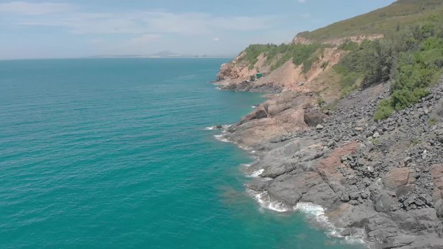 A flying camera takes a motorcyclist passing on a road near the sea with a view of the rocky shore.