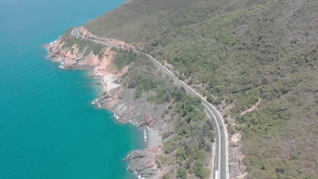 A flying camera takes a motorcyclist passing on a road near the sea with a view of the rocky shore.