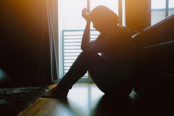 Asian woman sit on the floor beside window in dark room, place elbow on knees and other hand press...