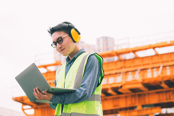 Image outside the industrial construction engineers in yellow protective ear muff discuss new project while using laptop and happy smile on the open building site near the crane.