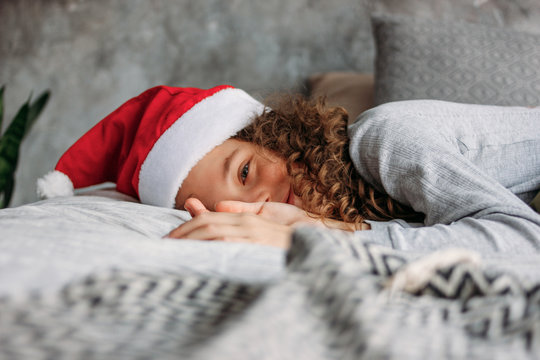 Curly haired beautiful tween girl in Santa hat and pajamas sitting on bed with pillow, christmas morning time