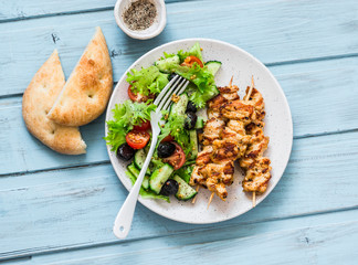 Chicken souvlaki kebabs, sun dried olives, tomatoes, cucumbers green salad and focaccia on blue background, top view