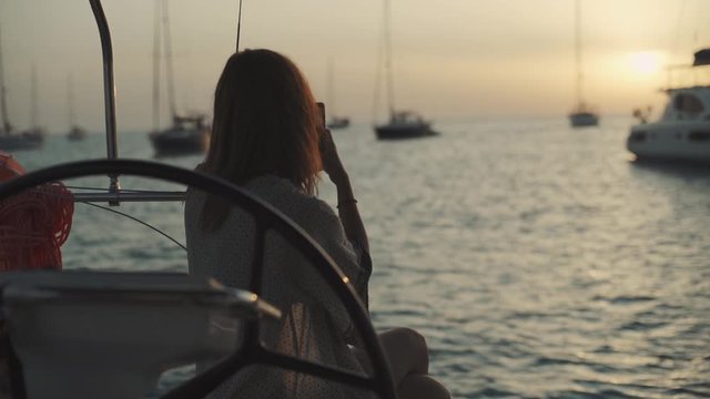 Beautiful woman on a yacht takes pictures on the phone of sunset on Cala Saona bay. Luxury yacht near the Formentera island