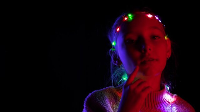 Adorable teenage girl with garland dancing in darkness. Portrait of cute happy child with illuminated garland on head and sweater dancing on black background