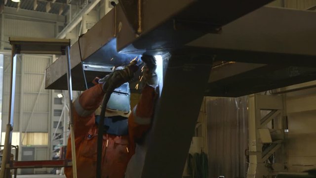 Steel worker welding on steel beam in factory hall- camera dolly movement - direction backward