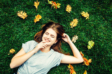 Happy 20 year old girl lies on green grass with autumn fallen leaves top view