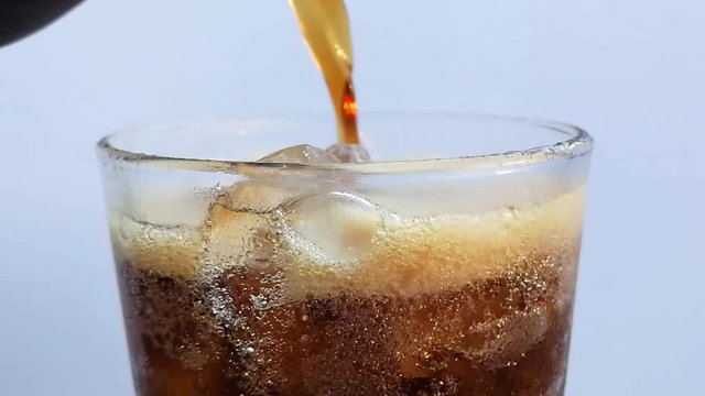 Pouring Cola to glass with ice and bubbles close-up. Zoom in glass of Cola fizzy drink over white background. Slow motion HD video footage.