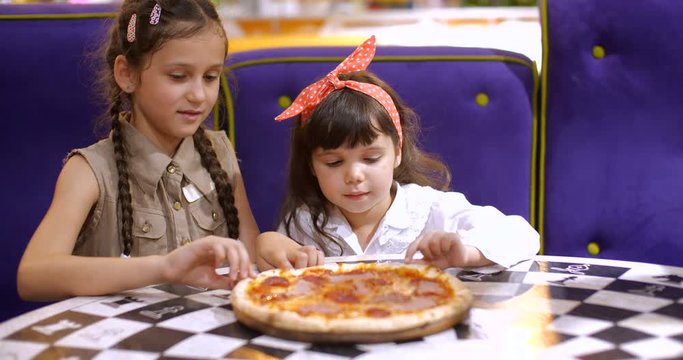Children eat pizza. Two cute girls of four and seven years old are sitting at a table in a pizzeria  taking slices of pizza and eating. Slow motion 50 FPS 4k