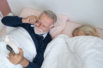 happy elderly couple caucasian senior man wake up and watching clock and old woman sleep together in white blanket in bedroom, retirement love lifestyle concept