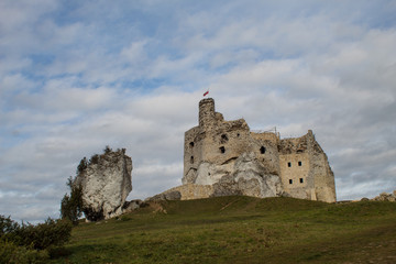 The ruins of a medieval European castle against the backdrop of a rocky ridge, blue sky, coniferous forest and green meadows.