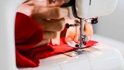 Hands of woman seamstress sewing red clothing on sewing machine with straight seam. She cuts...