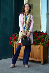 Full shot of brunette lady with a black handbag in a pink coat, blue trousers and gray shoes in...